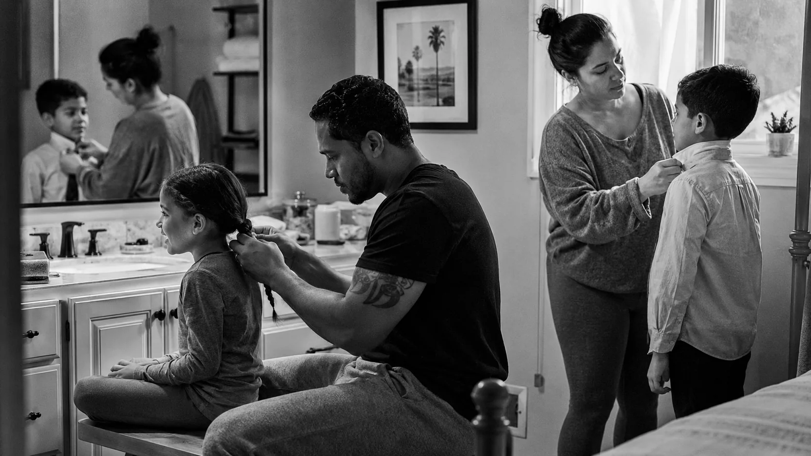 A father braids his daughter's hair at the bathroom counter while his son waits beside them
